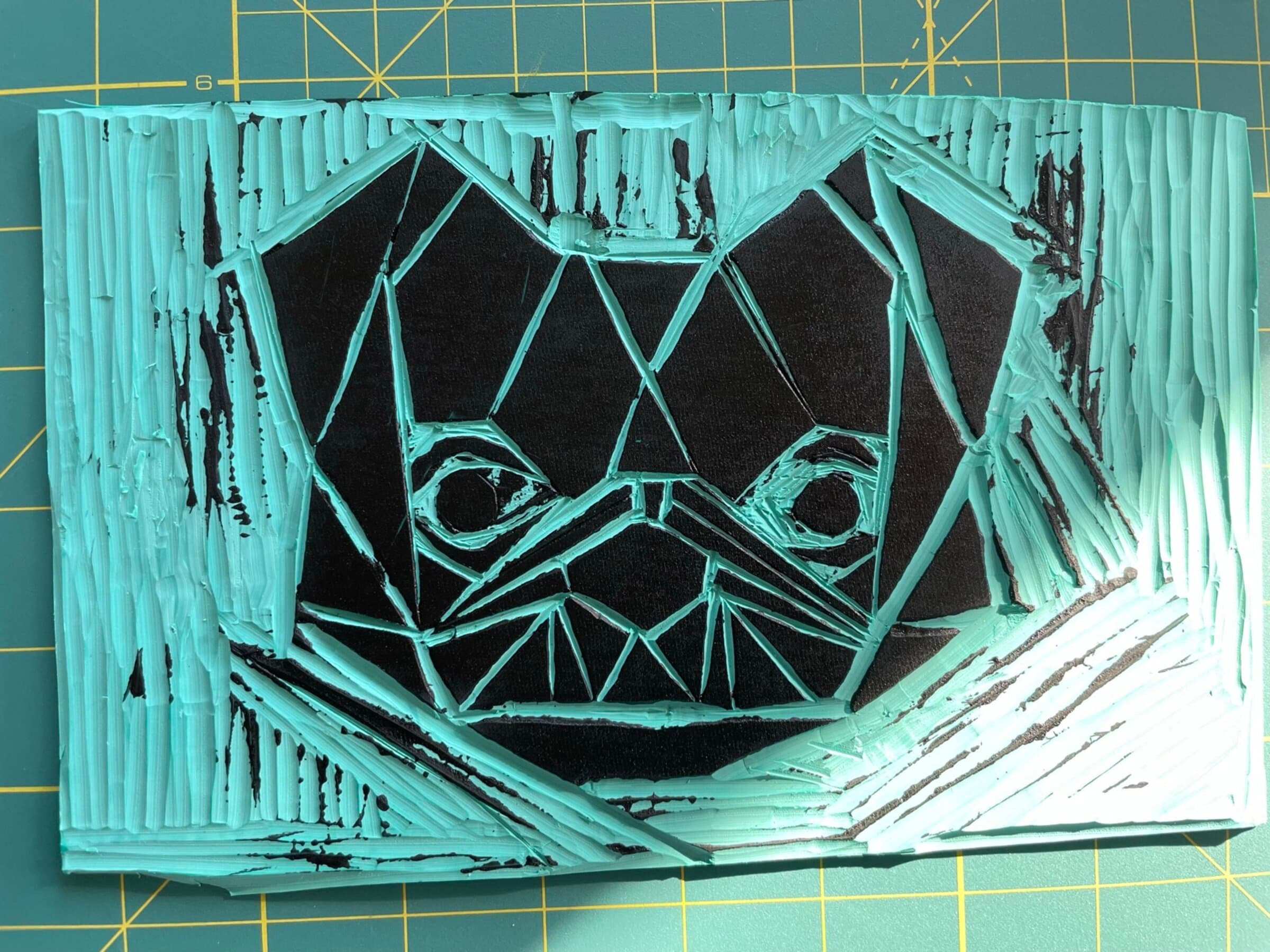 The carved bulldog block itself, photographed straight down on a green cutting mat. The lino is rolled with teal ink so the carved-out grooves read teal and the raised faces read black. The faceted-geometric composition of the dog's face is fully readable: planes for the head, triangles for the eyes, hatched grooves for the muzzle, scratch-textured fields surrounding the head.