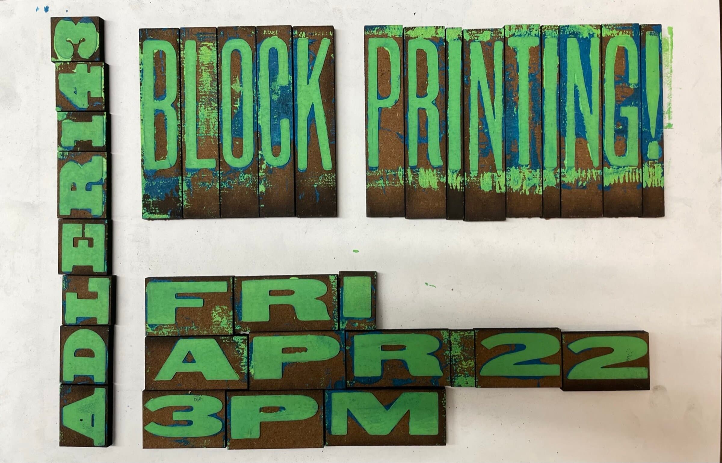The hand-carved wood letter blocks laid out on a white surface in the exact composition of the poster, each block inked in green: BLOCK PRINTING! across two horizontal rows in the upper third, AFTER143 set vertically along the left edge, FRI APR 22 3PM stacked underneath the title. Every letter still on its own individual wood block, the blue ink residue from a previous session visible on some block edges.