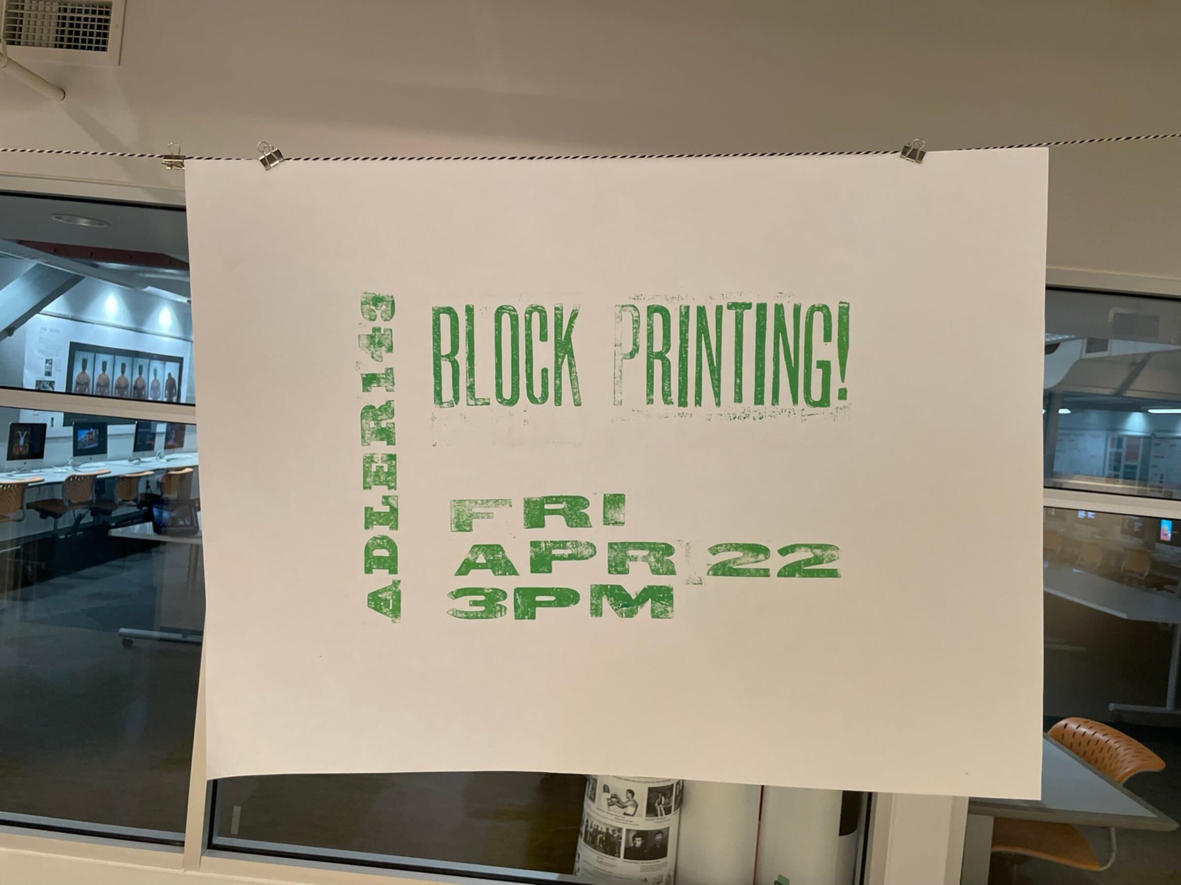 The same poster hanging from binder clips on a clothesline strung across a classroom-studio, large windows behind it, distressed green ink readable across the white sheet, classroom chairs and other prints visible through the windows in the background.