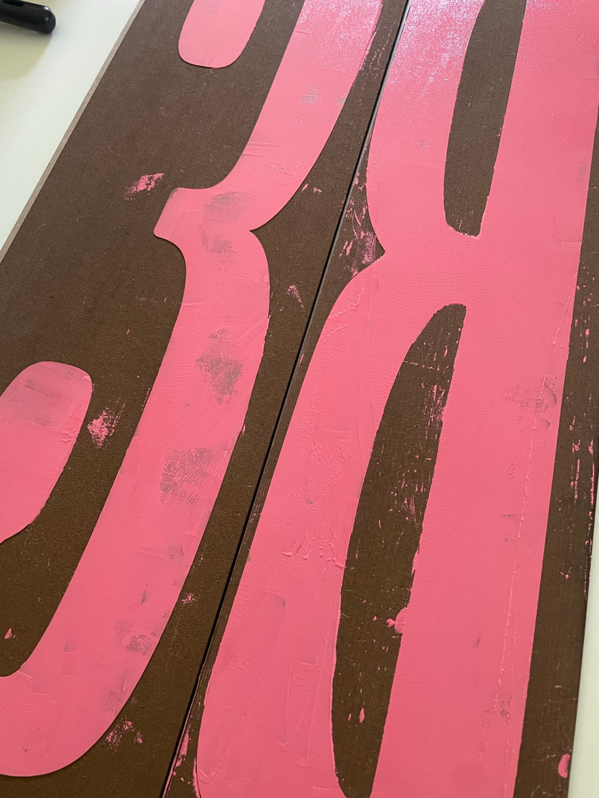 Two enormous wood planks side by side, each carved with one massive letter (a B and an S) and rolled with hot-pink ink. The carved-out areas read brown against the pink ink fields. Each plank is roughly the size of a coffee table, photographed at a raking angle so the textures of the inked surface catch the light.