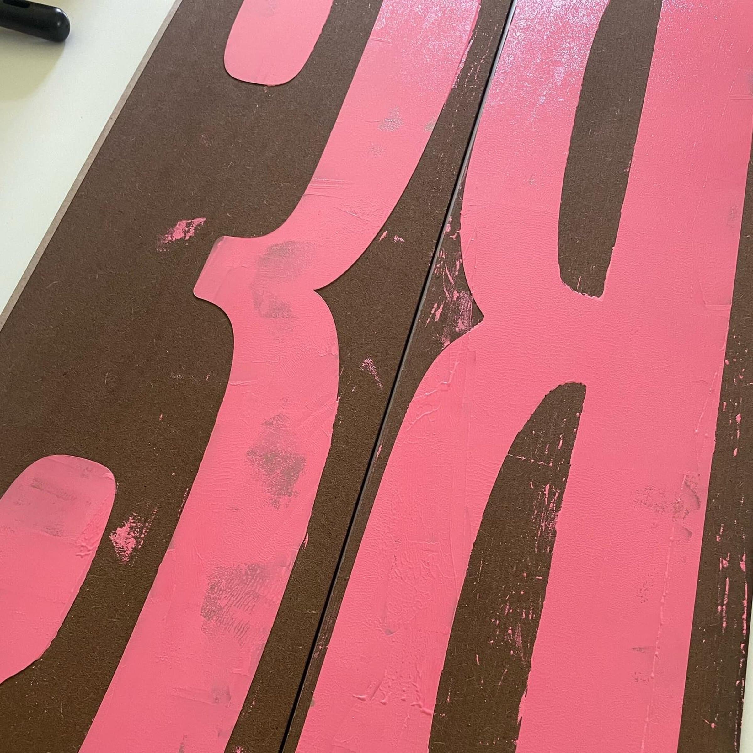 A tighter raking-angle close-up of the same two giant inked planks, the carved counters of the letters showing the brown wood underneath the pink ink. The seam where the two planks meet runs vertically through the middle of the frame.