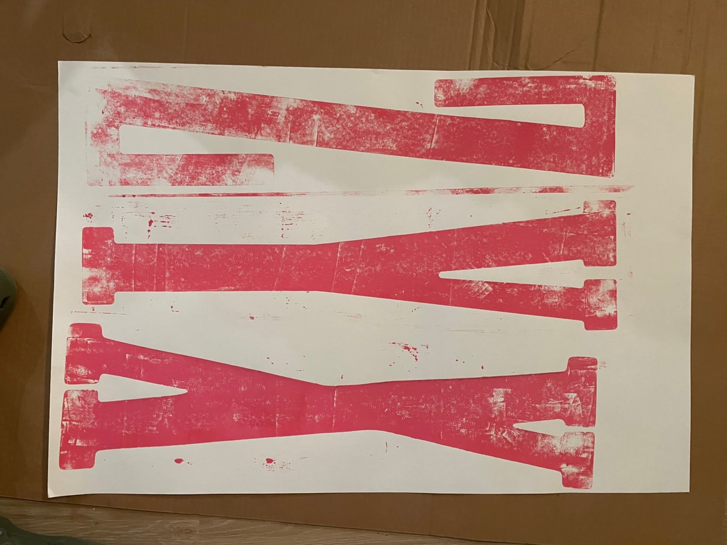 A second pull of three different giant carved letters (reading roughly K, K, I) printed in hot pink across a single white sheet on a cardboard floor. The sheet runs horizontal; each letter has its own band, the blocks separated by visible wood seams.