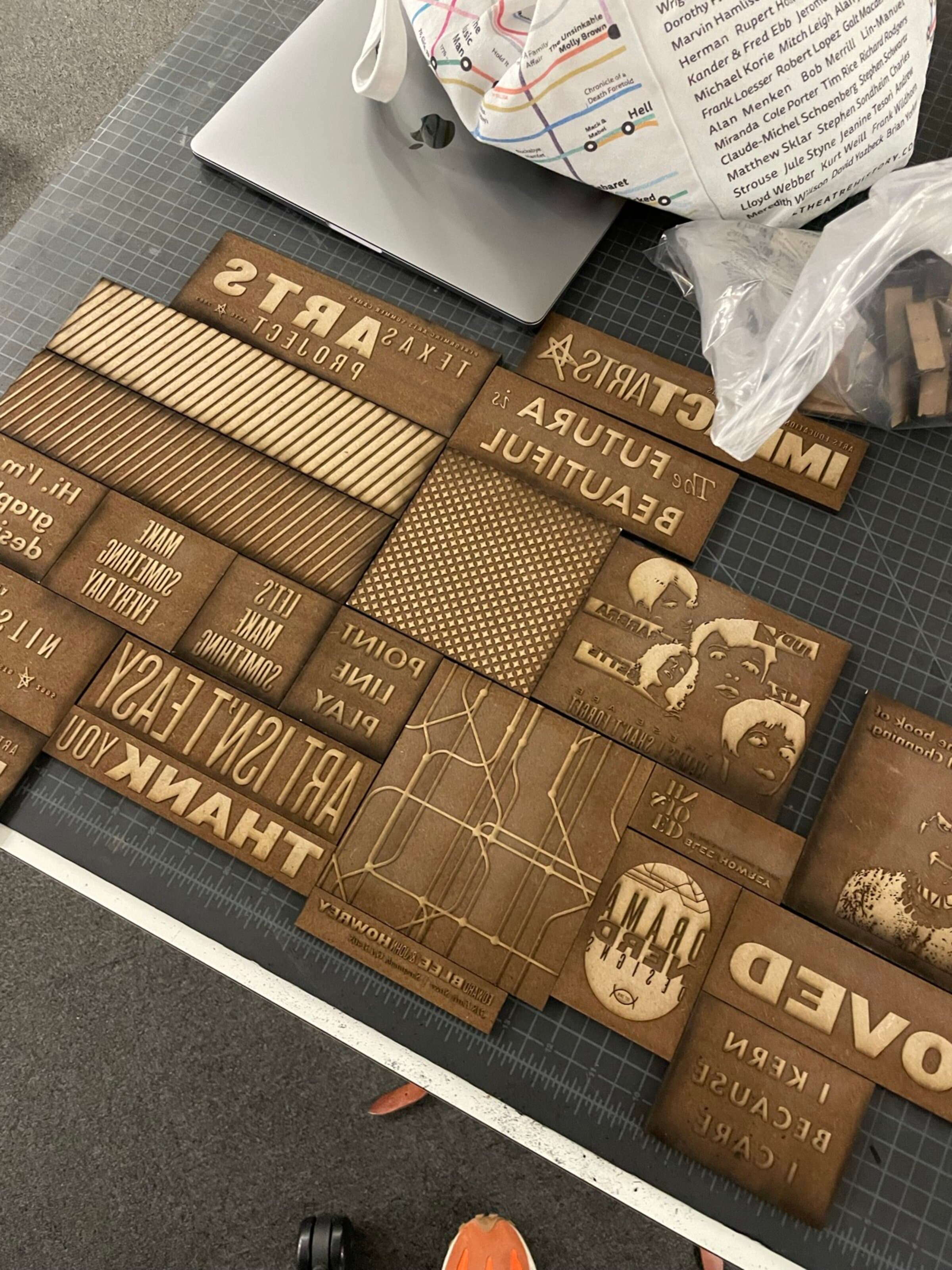 A massive flat-lay of laser-cut basswood letterpress blocks fitted edge-to-edge: phrase blocks reading ART ISN'T EASY, THANK YOU, MAKE SOMETHING, NEVER, FUTURA IS BEAUTIFUL, TEXAS, ARTS PROJECT, plus pattern blocks (vertical stripes, halftone dot field, a transit-map wireframe), a face block, a small label block. All set in mirror image so they print right-reading. The corner of an Apple laptop and a small subway-map sticker pack rest in the upper left for scale.
