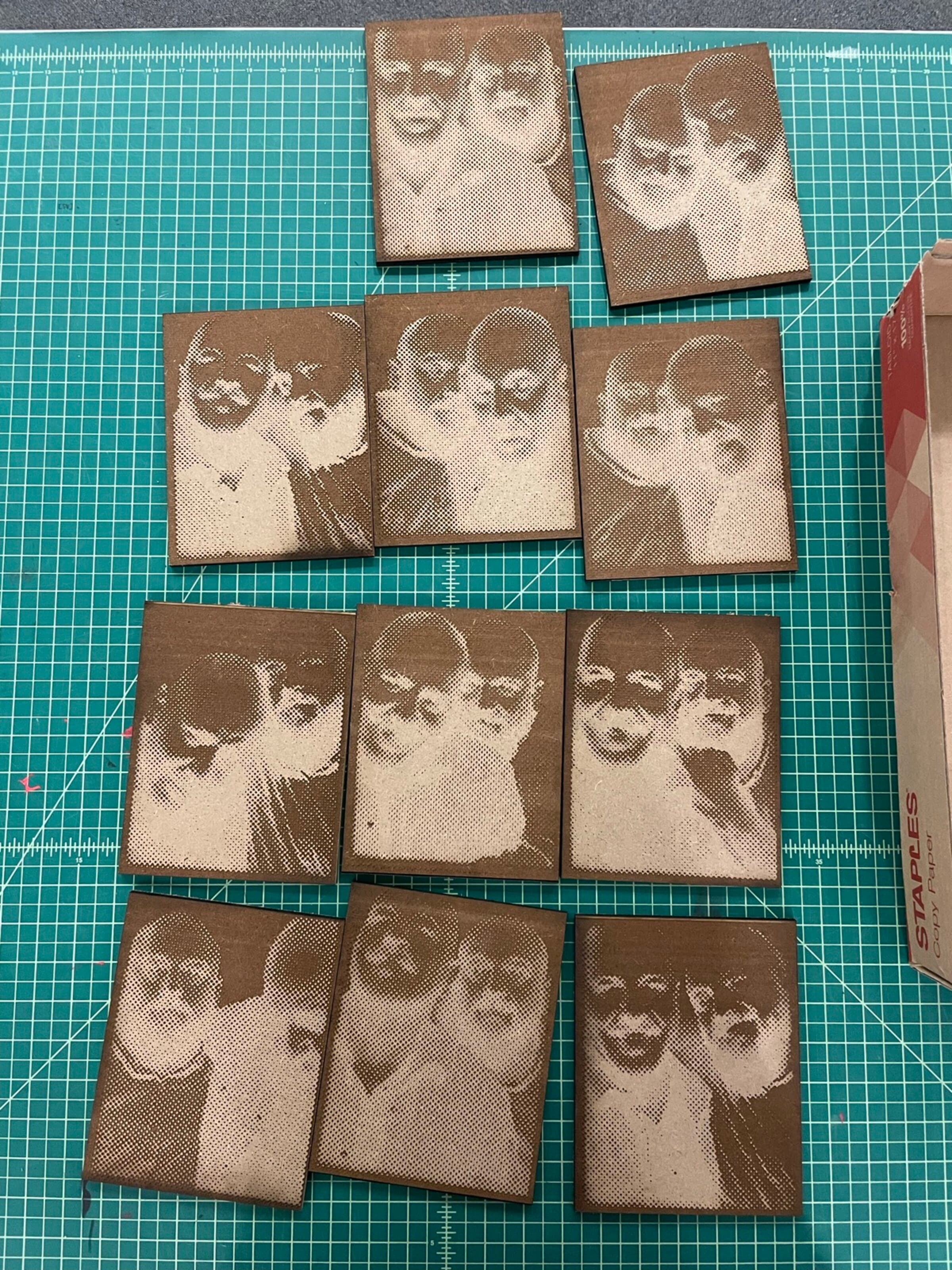 A flat-lay of about twelve small laser-engraved wood blocks tiled into a four-by-three grid on a green cutting mat. Each block carries the same multi-figure portrait composition burned into the wood, the engraving slightly varying in depth so the blocks read as a series. A 'STAPLES' box edge anchors the upper-right of the frame.