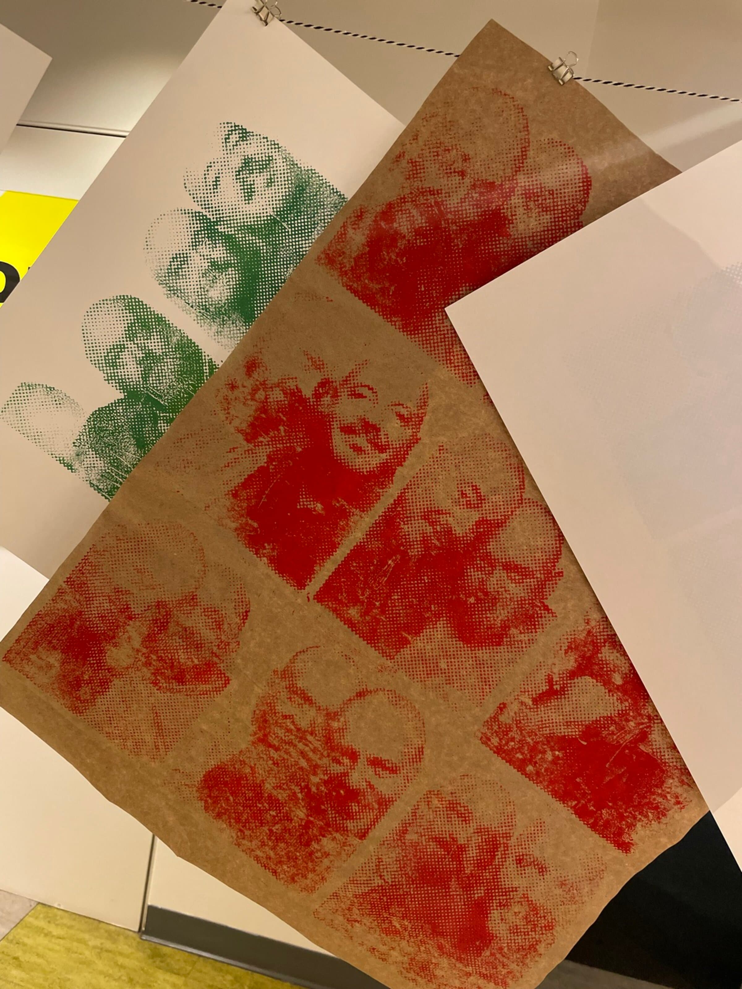 A working studio flat-lay: a kraft-paper sheet across the lower half of the frame stamped with a 3-by-3 grid of red multi-figure portraits, each one slightly off-register from the next. Below it, two prints in green halftone of the same composition lean against each other. A white reference sheet sits at the upper edge.