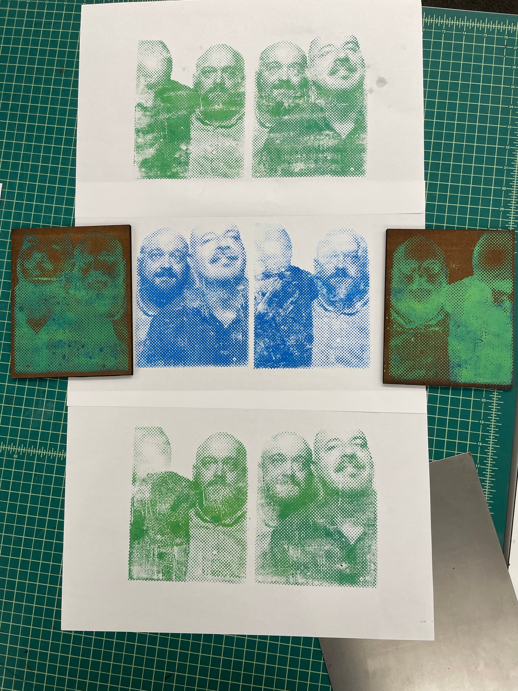 A flat-lay study of three side-by-side prints of the same multi-figure portrait, each pulled in a different ink: the left print in green, the center in cobalt blue, the right in a green-and-yellow split. Two reference blocks (also inked) sit at top and bottom of the spread on a green cutting mat.