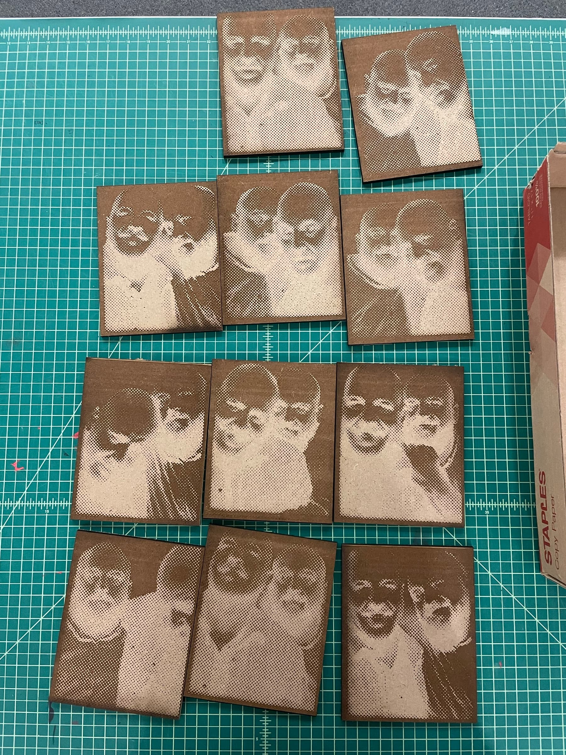 A flat-lay grid of about a dozen identical halftone-style portrait prints lined up across a green cutting mat. Each is the same multi-figure composition pulled in slightly different ink loads of brown and sepia, the cardboard backings catching warm directional light.