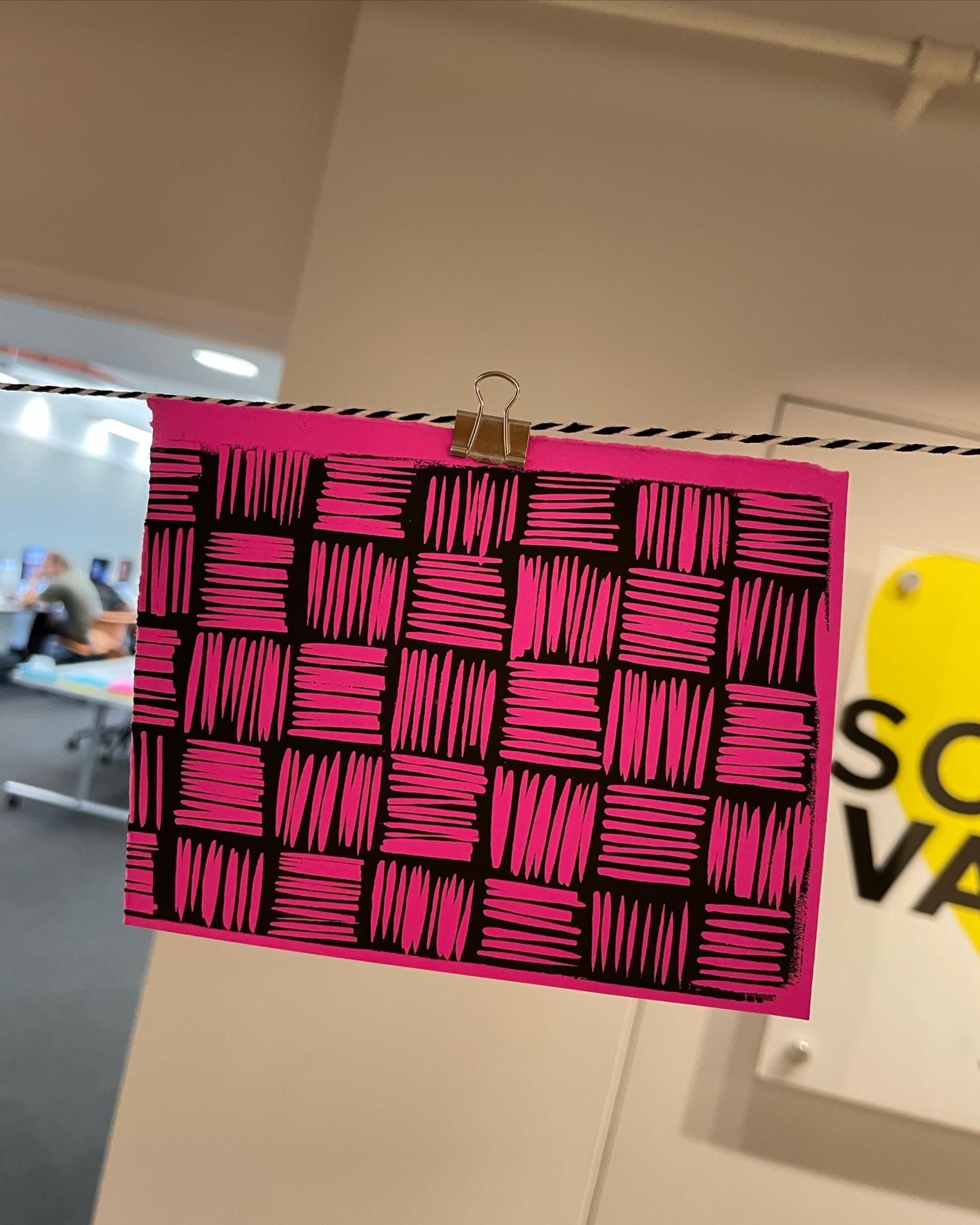 The same hatched-checkerboard print pinned to a clothesline in a different studio space, a yellow SoVA poster visible on the wall behind, classroom desks and chairs visible in the deeper background. The print catches directional light from the right.