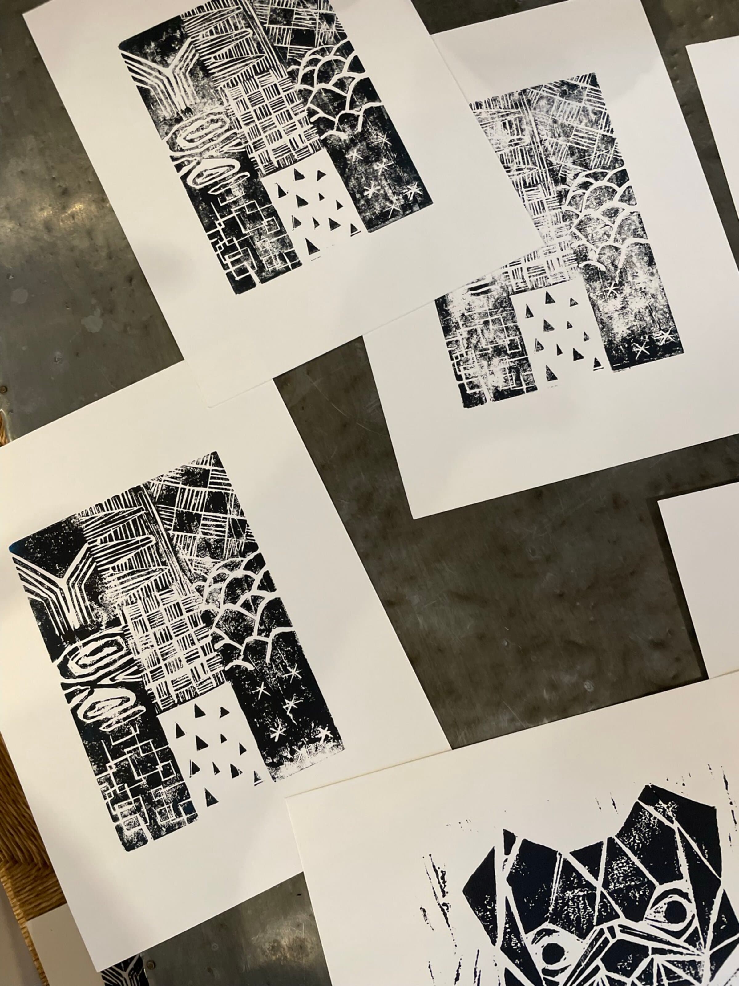 A studio flat-lay on a metal worktable: three pulls of the texture-grid block printed in black ink across white paper, the prints overlapping each other as they dry. A black halftone face print (the bulldog) leans against the upper edge of the frame as a reference pull from the same session.