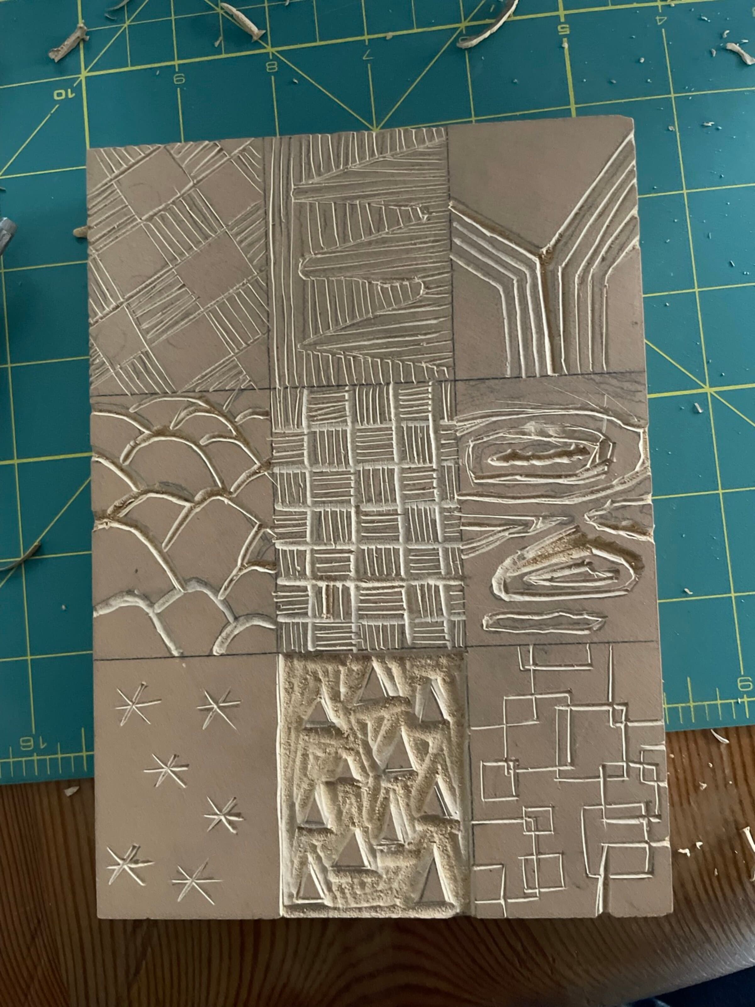 A fresh-carved 9-up grid of texture studies cut into linoleum, photographed straight down on a green cutting mat. Uninked: the carved-out grooves catching shadow against the raw lino. Patterns include chevrons, OK lettering rough, a woven plaid, dragon scales, geometric brick, snowflakes, and irregular geometric shards. Carving curls of waste linoleum scattered around the edges of the mat.