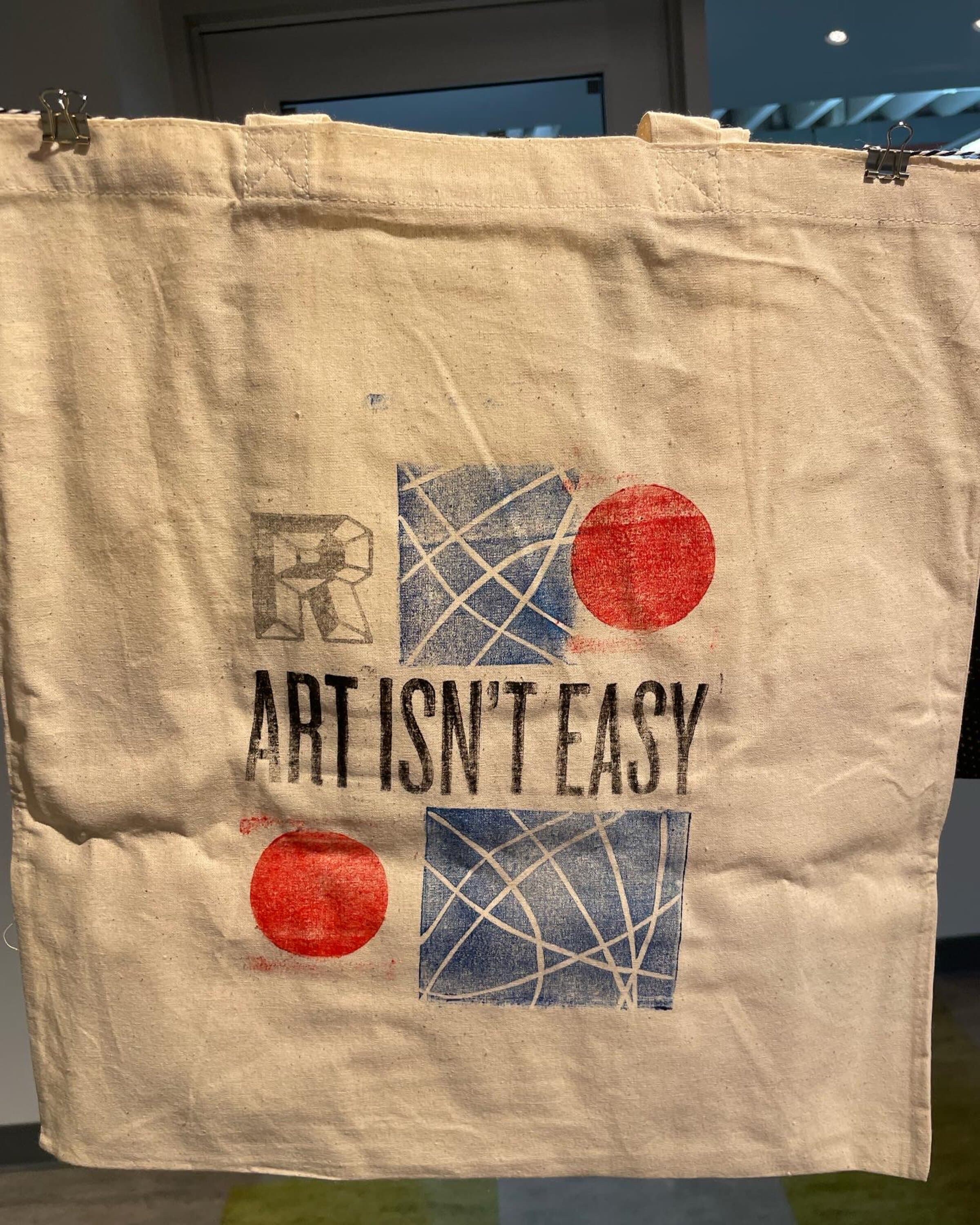 A second canvas tote in an alternate composition: ART ISN'T EASY set in chunky distressed black display caps anchored low-center, an R letter block in the upper-left, two indigo bramble-line blocks stacked on the right, and two scarlet ink discs at upper-right and lower-left. The same elements rearranged into a different layout.