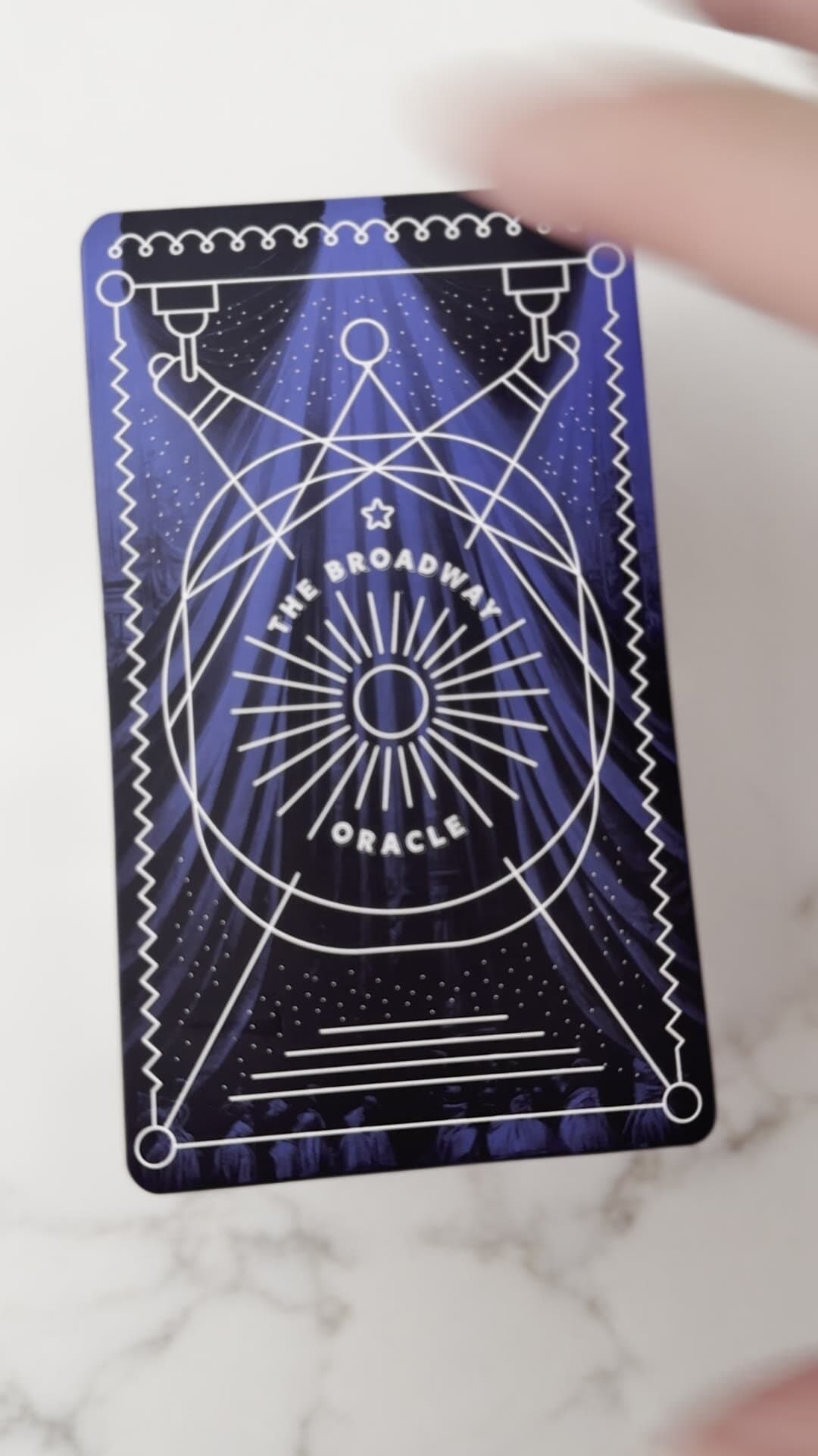 A single printed card held to the camera: same composition rendered in white-on-royal-blue, the curtain folds painted into the negative space behind the spotlights, the sun-ray oracle emblem centered, the dimensions and proportions of an actual playing card.