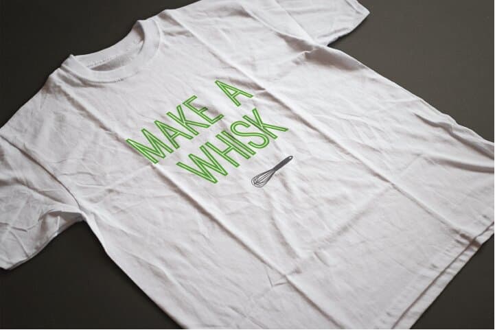 A white t-shirt printed with MAKE A WHISK in green outline display caps, set with a small whisk icon as the punctuation. Photographed flat on a charcoal surface.