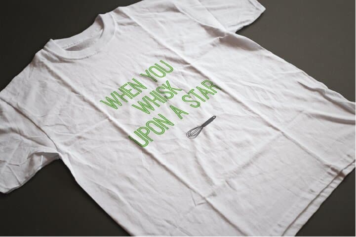 A white t-shirt printed with WHEN YOU WHISK UPON A STAR in green outline display caps, the whisk icon to the right of the line, photographed flat at an angle on a charcoal surface.