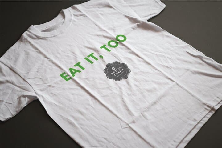 A white t-shirt printed with EAT IT, TOO set in solid green caps, paired with the small Buttercream badge icon. Photographed flat on a charcoal surface.