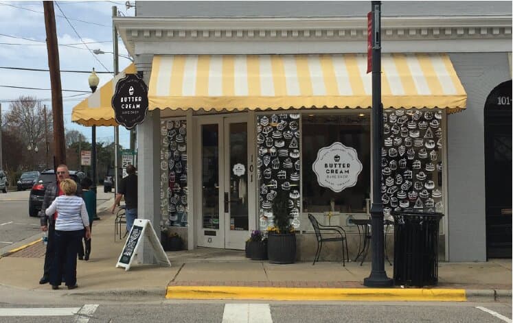 The actual storefront on a cooler day: the same yellow awning and white window caps, with the cupcake-pattern decals visible on the side windows wrapping around the BUTTERCREAM BAKE SHOP centered medallion.