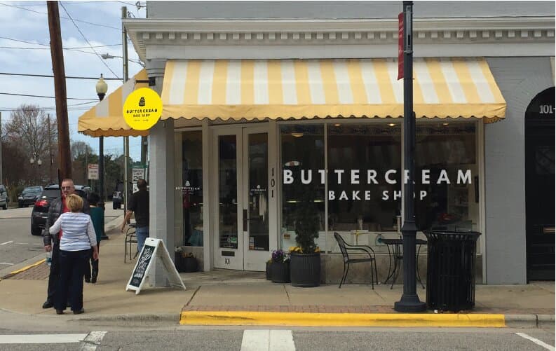 The actual storefront from across the street: yellow-and-cream striped awning, BUTTERCREAM set in white caps across the picture window with BAKE SHOP underneath, the round badge sign on the awning bracket, sidewalk seating, customers walking up.