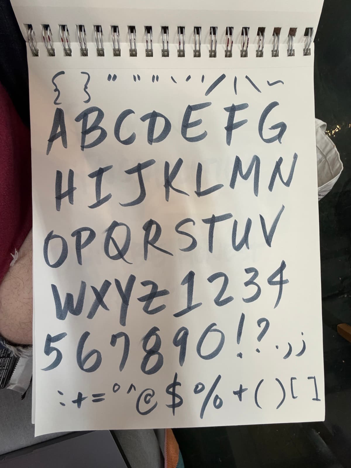 The original drawn alphabet on a Strathmore spiral pad: a complete uppercase A through Z plus numerals and punctuation, hand-lettered in black felt-tip on white paper, the whole thing photographed flat with the spiral binding visible on the left edge