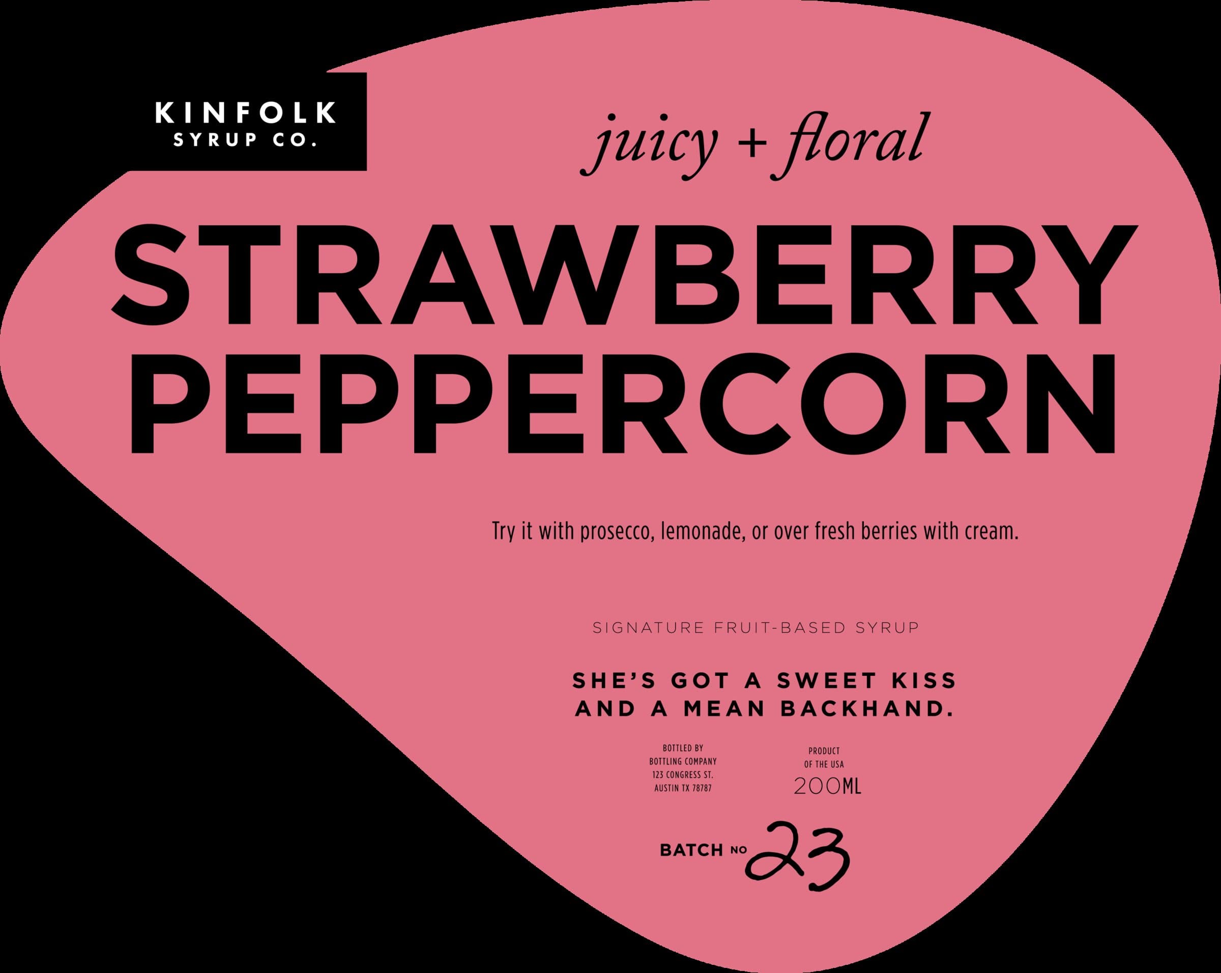 The Strawberry Peppercorn label: hot dusty-pink teardrop, KINFOLK SYRUP CO. tag in the upper-left, 'juicy + floral' italic kicker, STRAWBERRY PEPPERCORN in chunky black display caps, 'Try it with prosecco, lemonade, or over fresh berries with cream' as the serving suggestion, and 'She's got a sweet kiss and a mean backhand' as the personality line.