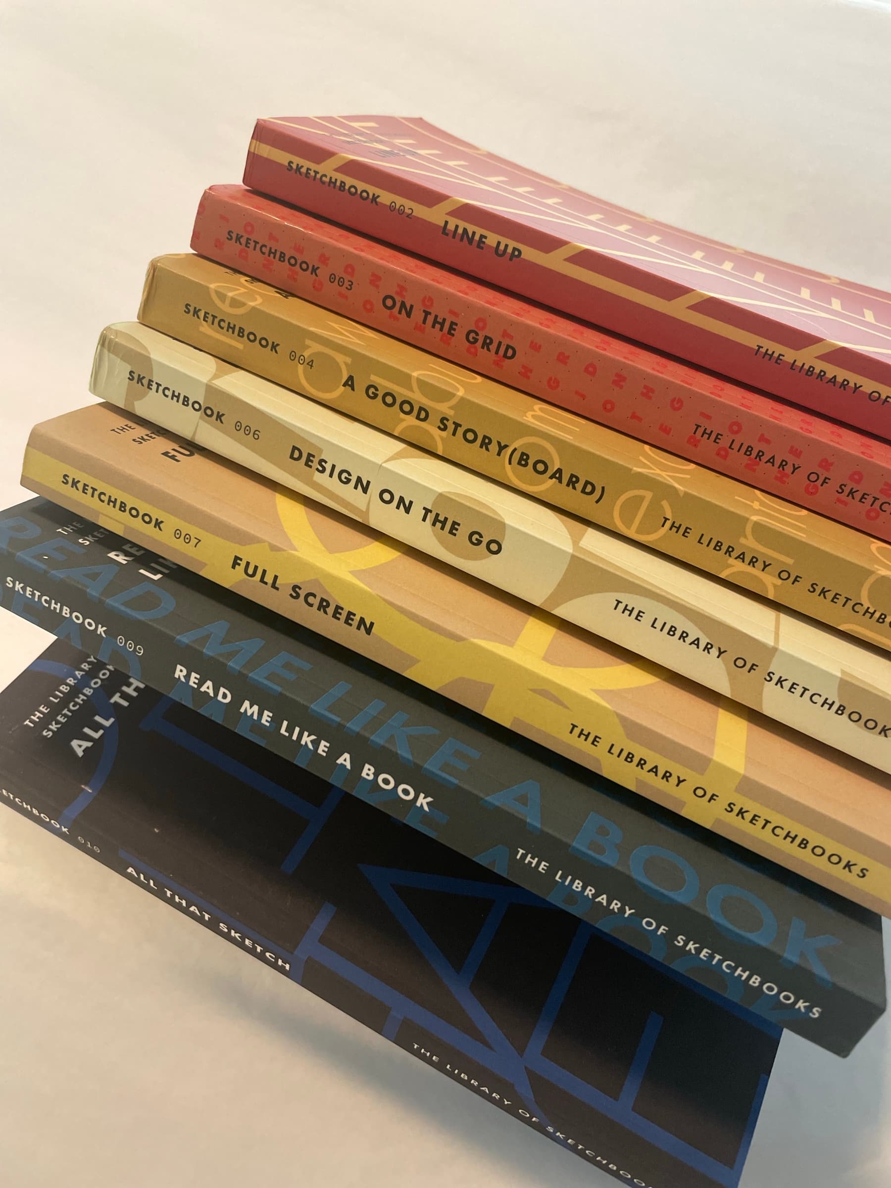 The set fanned out at an angle: nine of the ten spines visible, each volume number and title legible, the pattern detail of every book different, the LIBRARY OF SKETCHBOOKS runner running uniformly across them all.