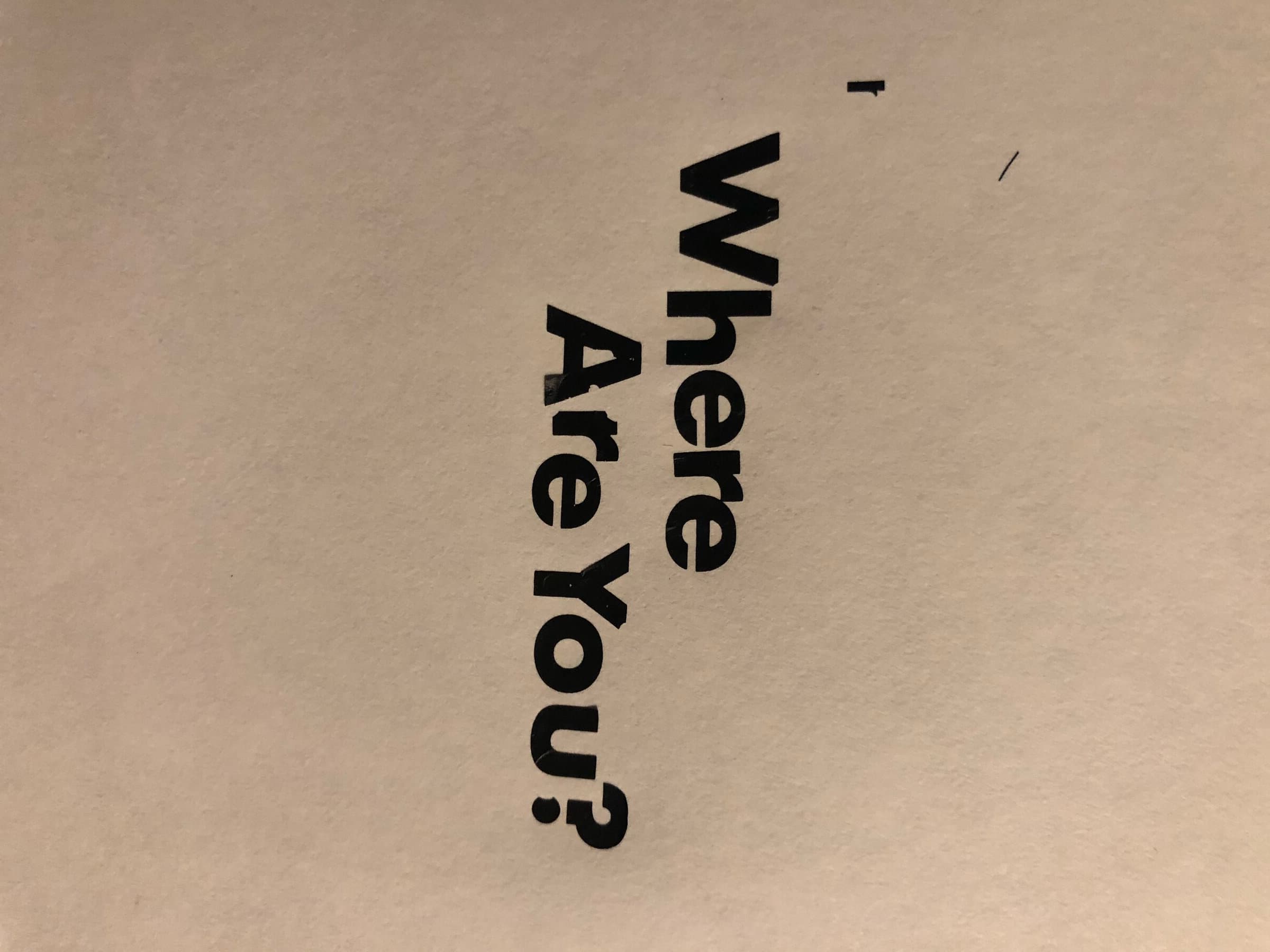 A blank sheet of paper with WHERE ARE YOU? hand-set vertically in two stacked lines of chunky black display caps, photographed at an angle on a wood-grain surface.