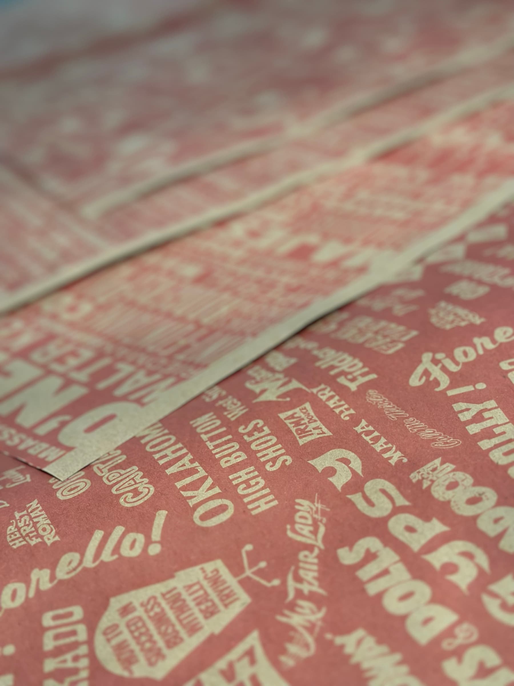 A close-up of the alphabet wrapping paper layered as multiple sheets: cream show titles like Fiorello!, Oklahoma!, High Button Shoes, My Fair Lady, Hot Mikado, Mata Hari, and Captain set in mixed display caps, italics, and scripts at multiple sizes against the coral kraft ground.